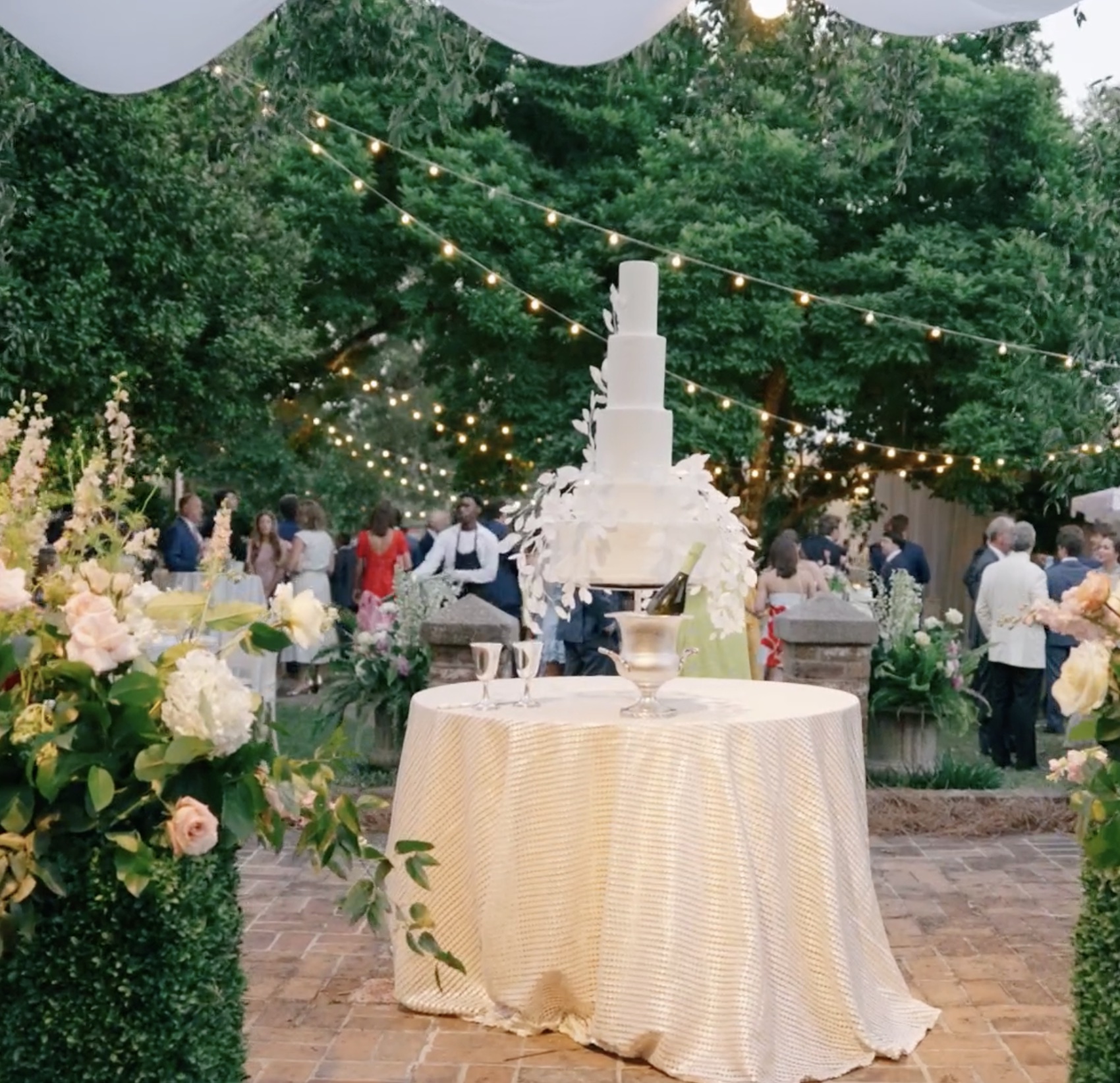 Four-tier wedding cake on gold linen with string lights and guests at dusk