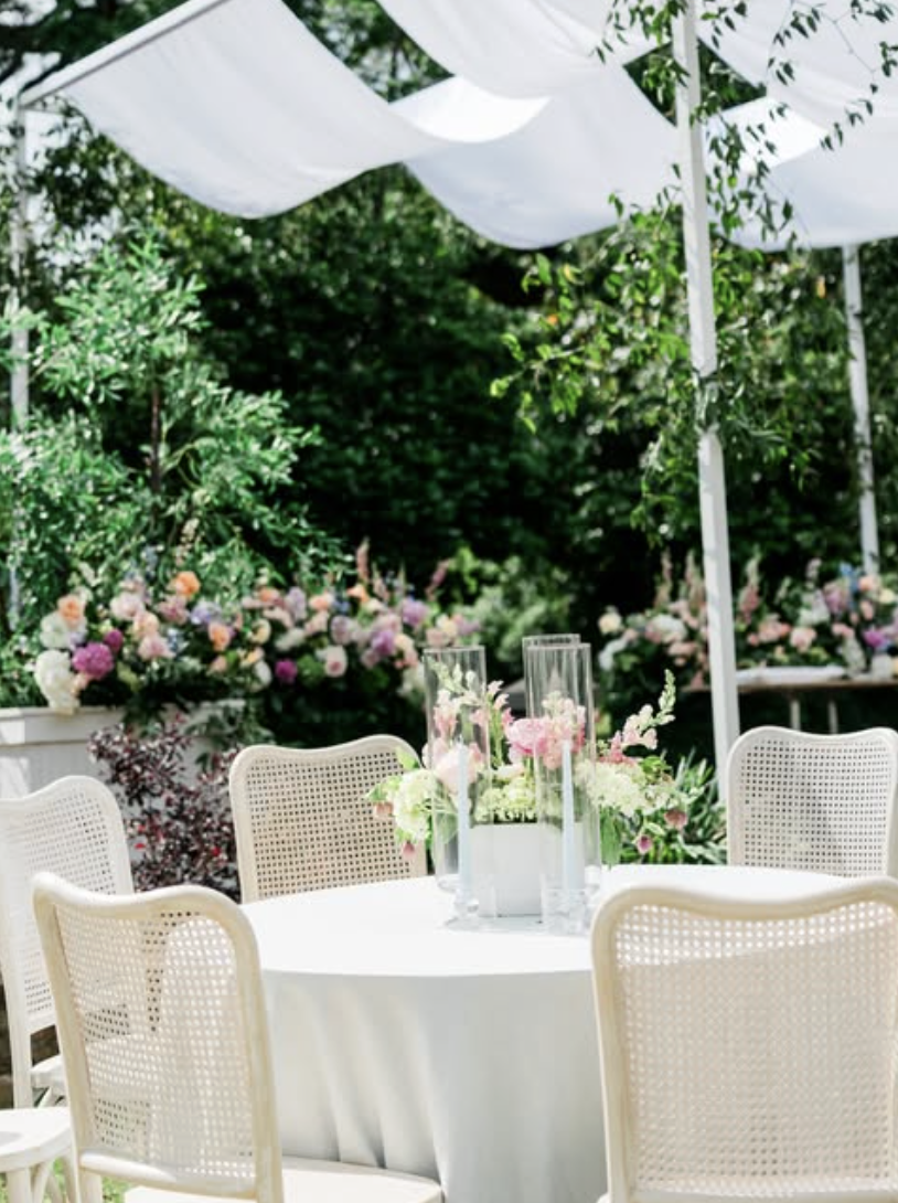 White cane-back chairs around round table with pink floral centerpiece and glass candles in garden setting