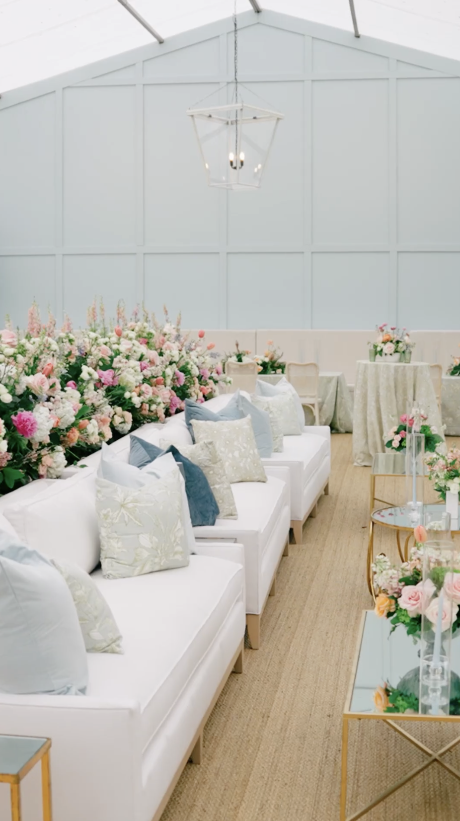 White lounge sofas with blue and sage pillows backed by a lush floral wall under a tent